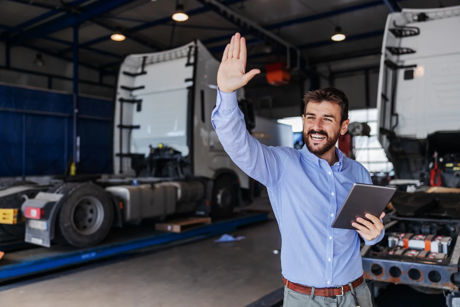 A smiling depot manager holding a tablet and waving in a truck garage.