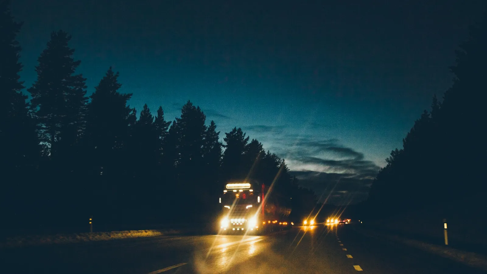 A work truck with its lights on coming down a forest highway at dusk.